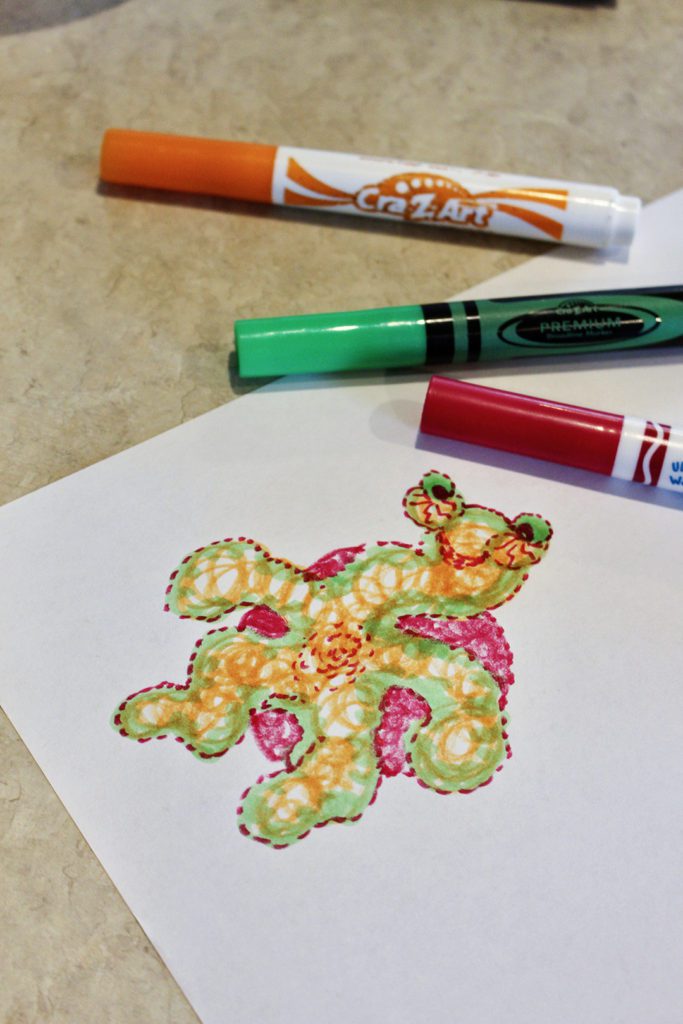 An orange and green scribble monster colored on a piece of paper with markers to the side.
