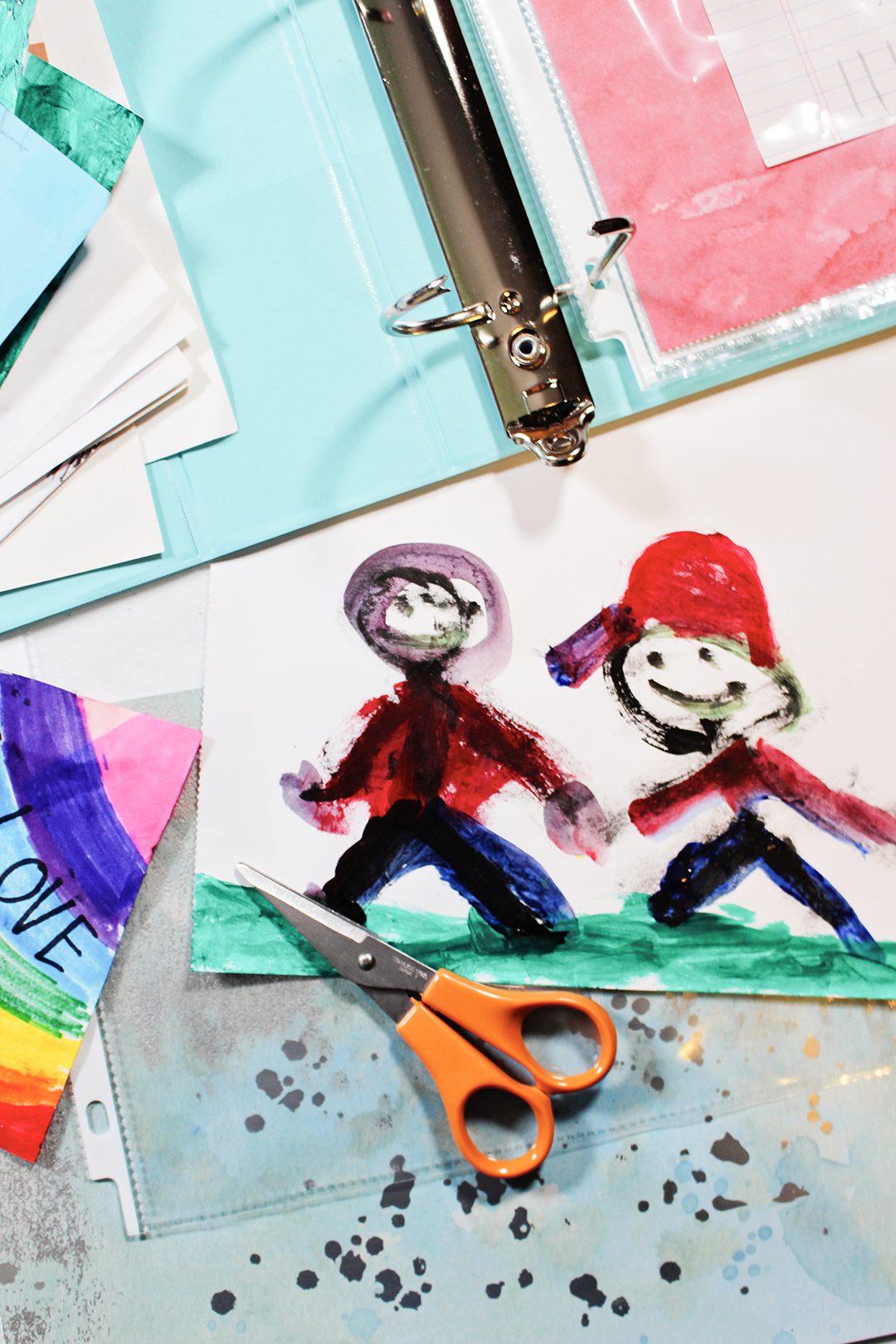 A child's painting of two people holding hands, with scissors, a binder and scrapbook paper nearby.