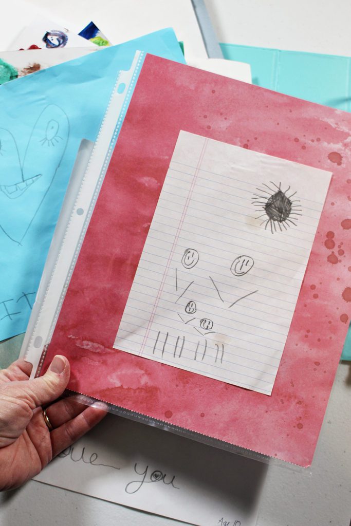 A child's pencil drawing of a family and sun, mounted on scrapbook paper and inside of a sheet protector.