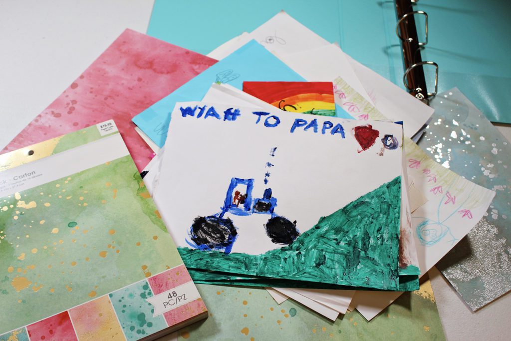 A child's painting of a tractor and a hill, addressed to Papa, scrapbook paper and a binder in the background.