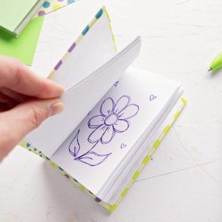 A DIY miniature journal with a flower picture drawn on one of the pages, scrapbook paper and pens in the background.