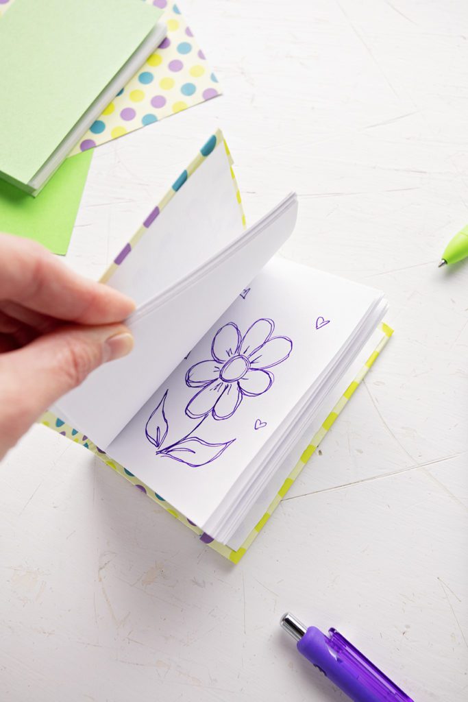 Un journal miniature DIY avec une image de fleur dessinée sur l'une des pages, du papier de scrapbooking et des stylos en arrière-plan.