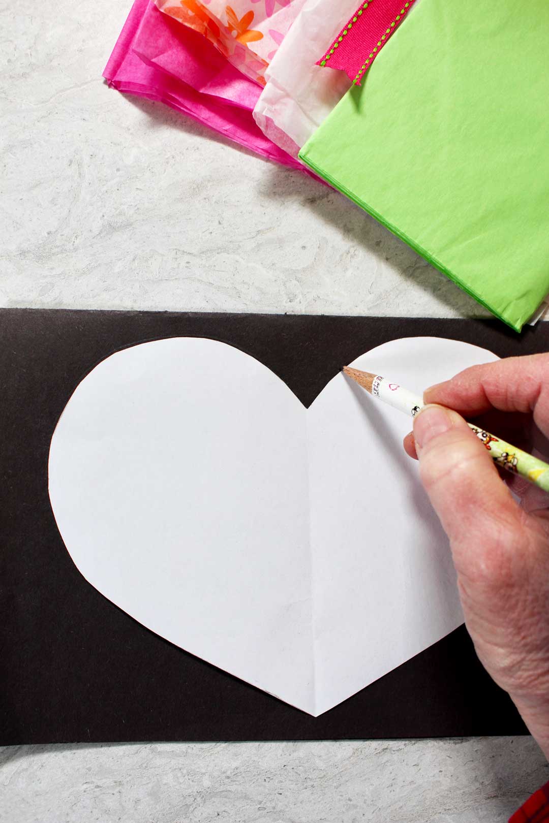 Pink, green and flower patterned tissue paper near a pencil outlining a heart on a black piece of paper.