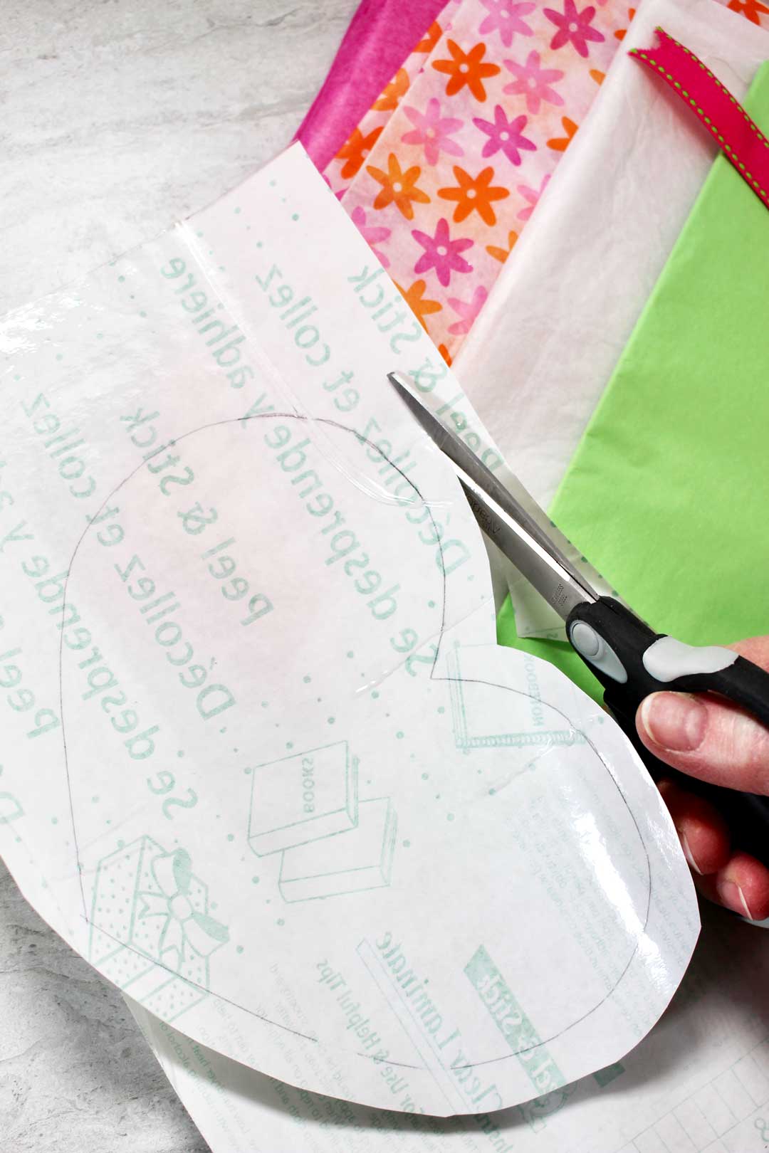 Cutting a heart pattern from contact paper, near pink, green, and flower patterned tissue paper.