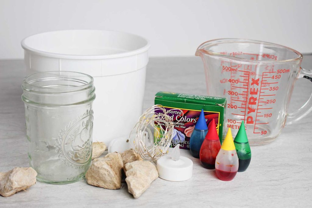 A bucket of water, a mason jar, rocks, food coloring, a glass measuring cup, and a tea light candle.