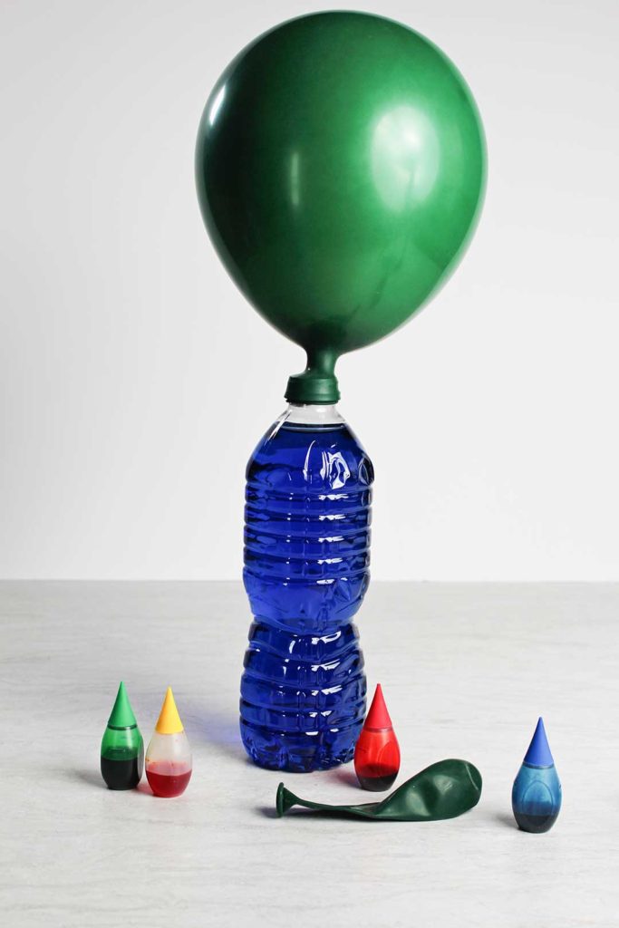 A green balloon filled with air attached to the top of a water bottle filled with blue water, surrounded by food coloring bottles.