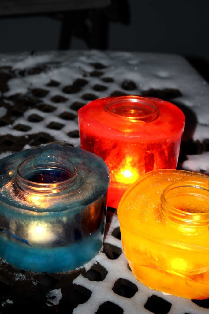 Yellow, blue, and red frozen luminaries with candles lit inside, sitting on a table with snow outside.
