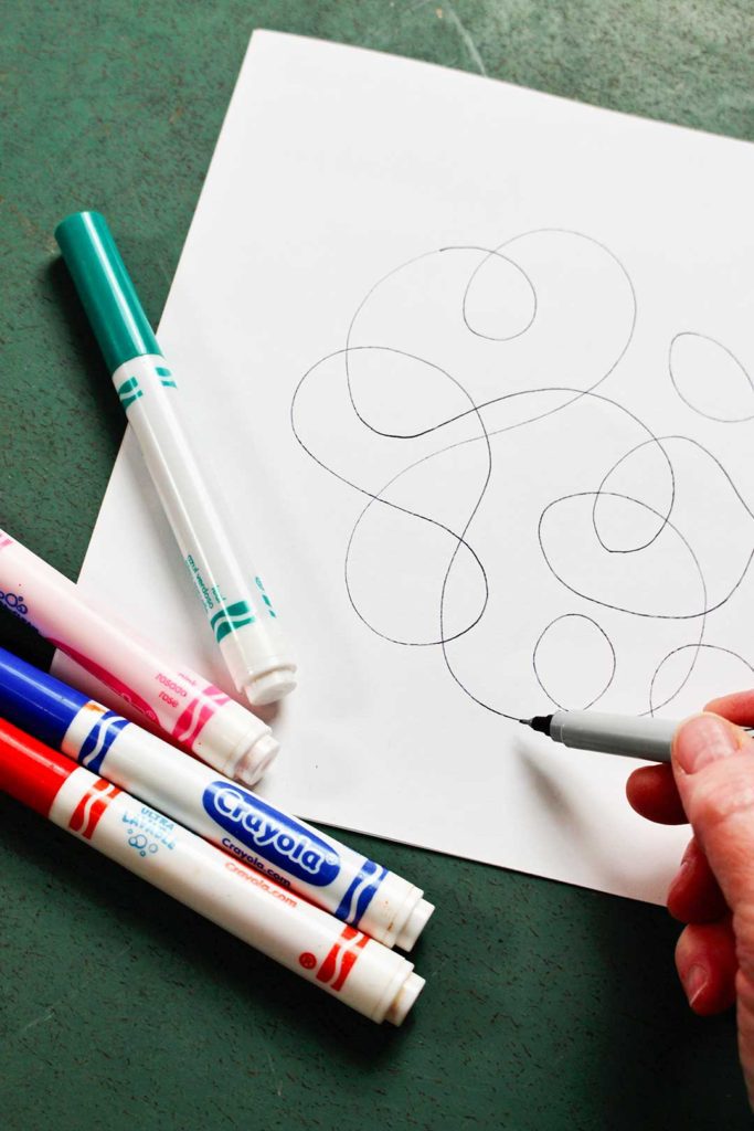 Using a marker to draw scribbles around a piece of white paper, surrounded by colorful markers.