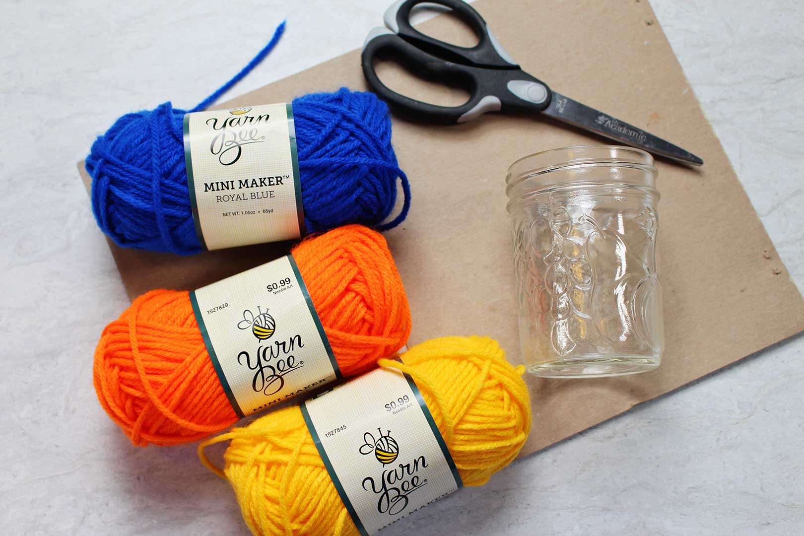 Blue, orange, and yellow yarn near a pair of scissors, jar, and piece of cardboard.