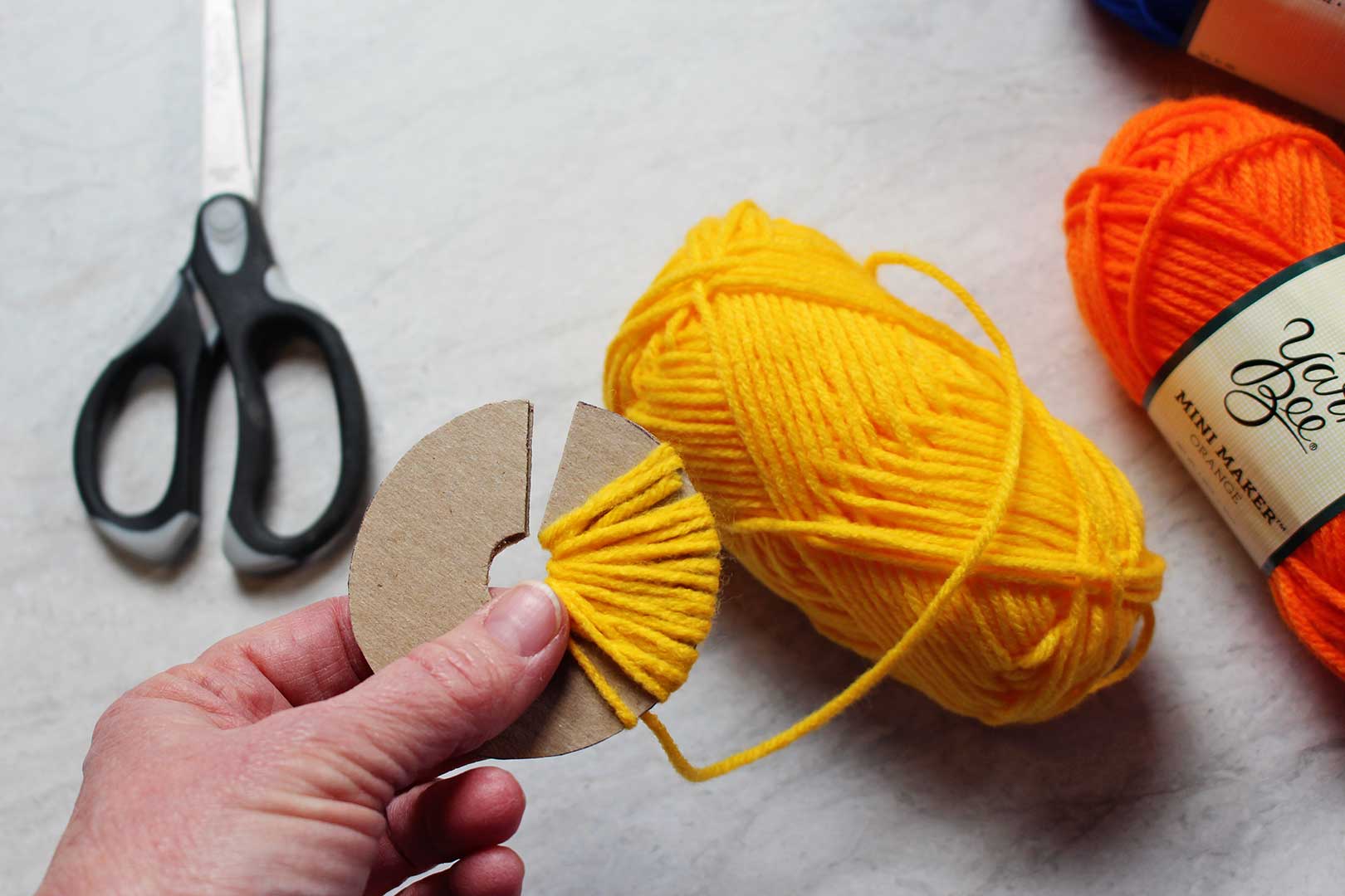 Yellow yarn wrapped around a circular cardboard shape, scissors and orange yarn in the background.