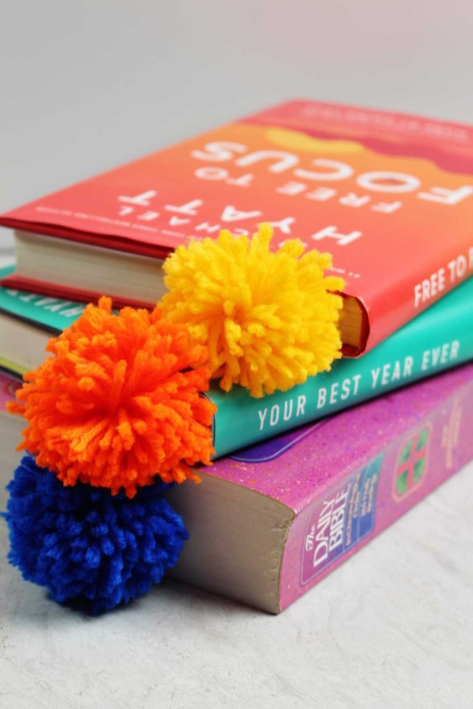 A yellow, blue and orange pom pom bookmark inside a stack of books.