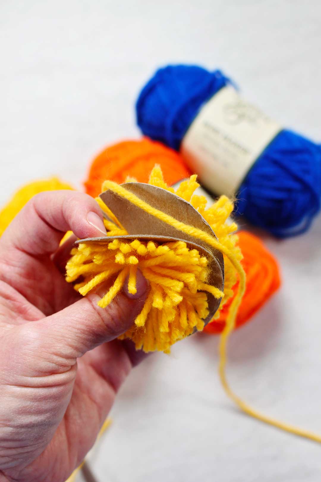 Yellow yarn cut around a cardboard circle to create a pom pom, blue and orange yarn in the background.