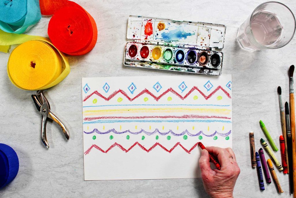 Coloring zigzag lines with a red crayon, surrounded by watercolor paints, paintbrushes, and streamers.