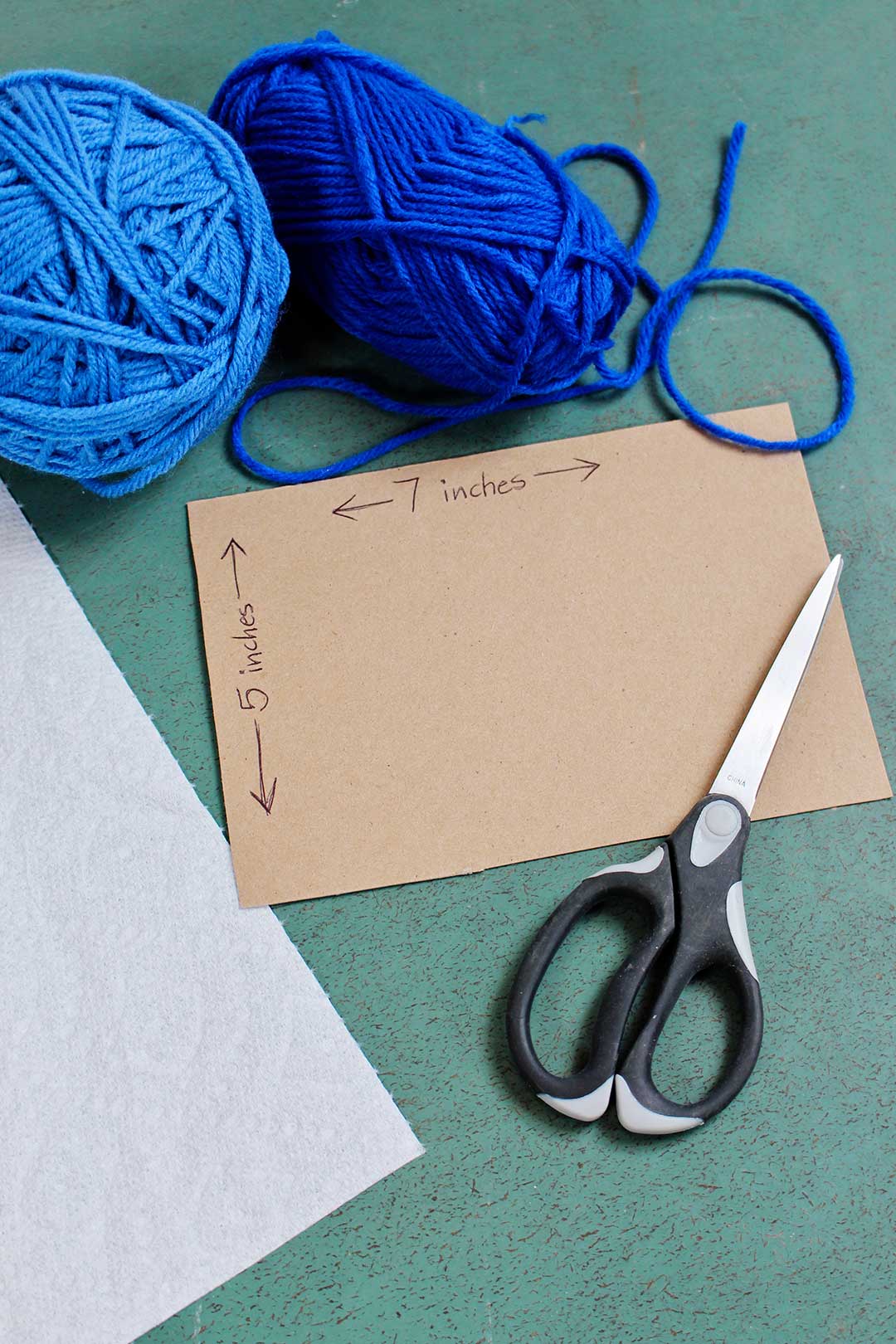 A 5x7 inch piece of cardboard surrounded by a pair of scissors, paper towel, and blue yarn.