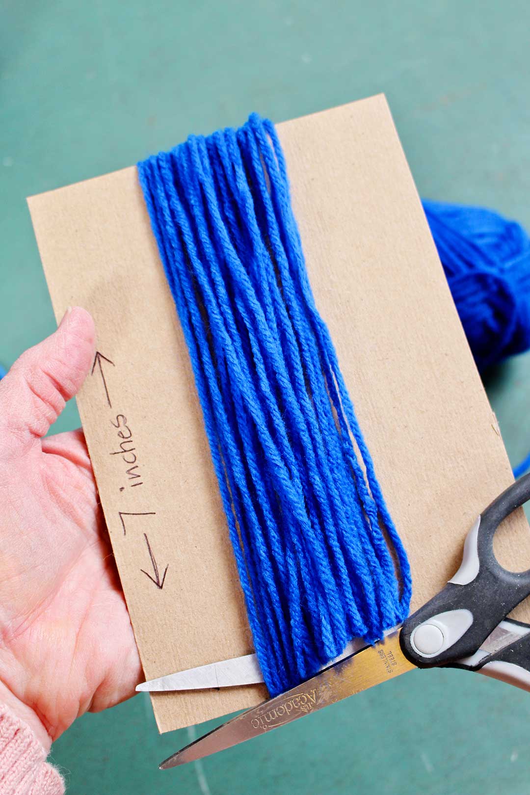 Blue yarn wrapped around a piece of cardboard, being cut by a pair of scissors.