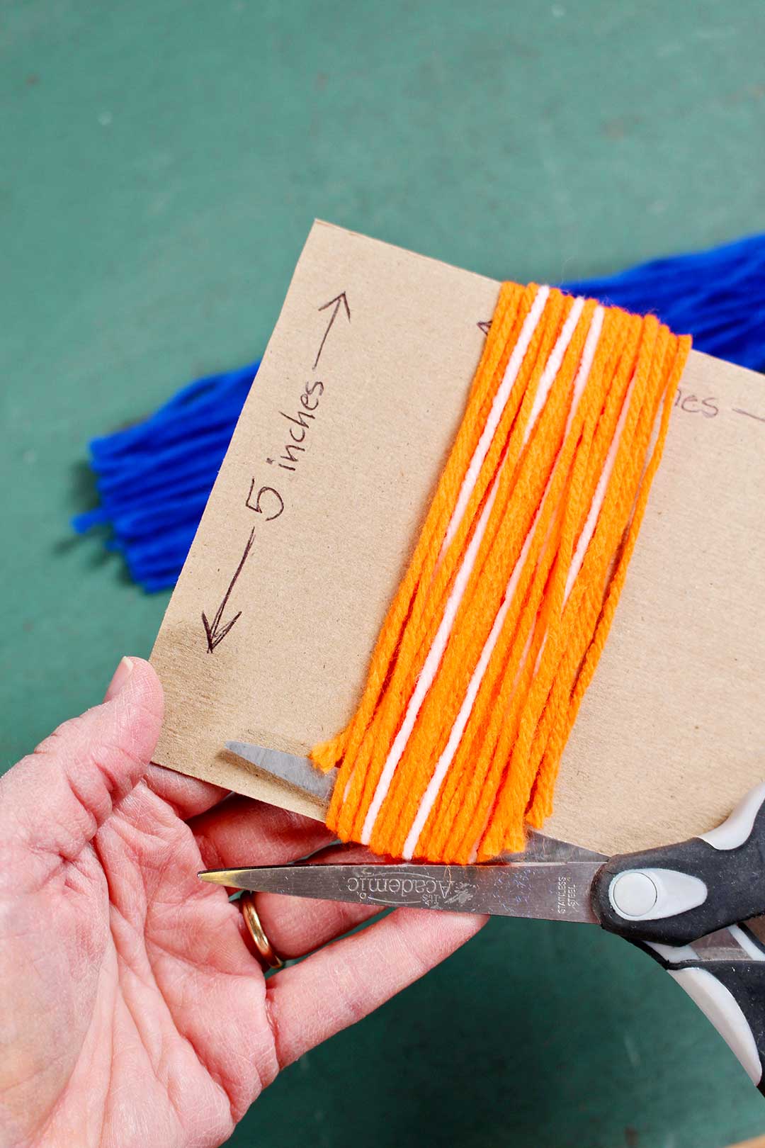 Orange and white yarn wrapped around a piece of cardboard, being cut by a pair of scissors.