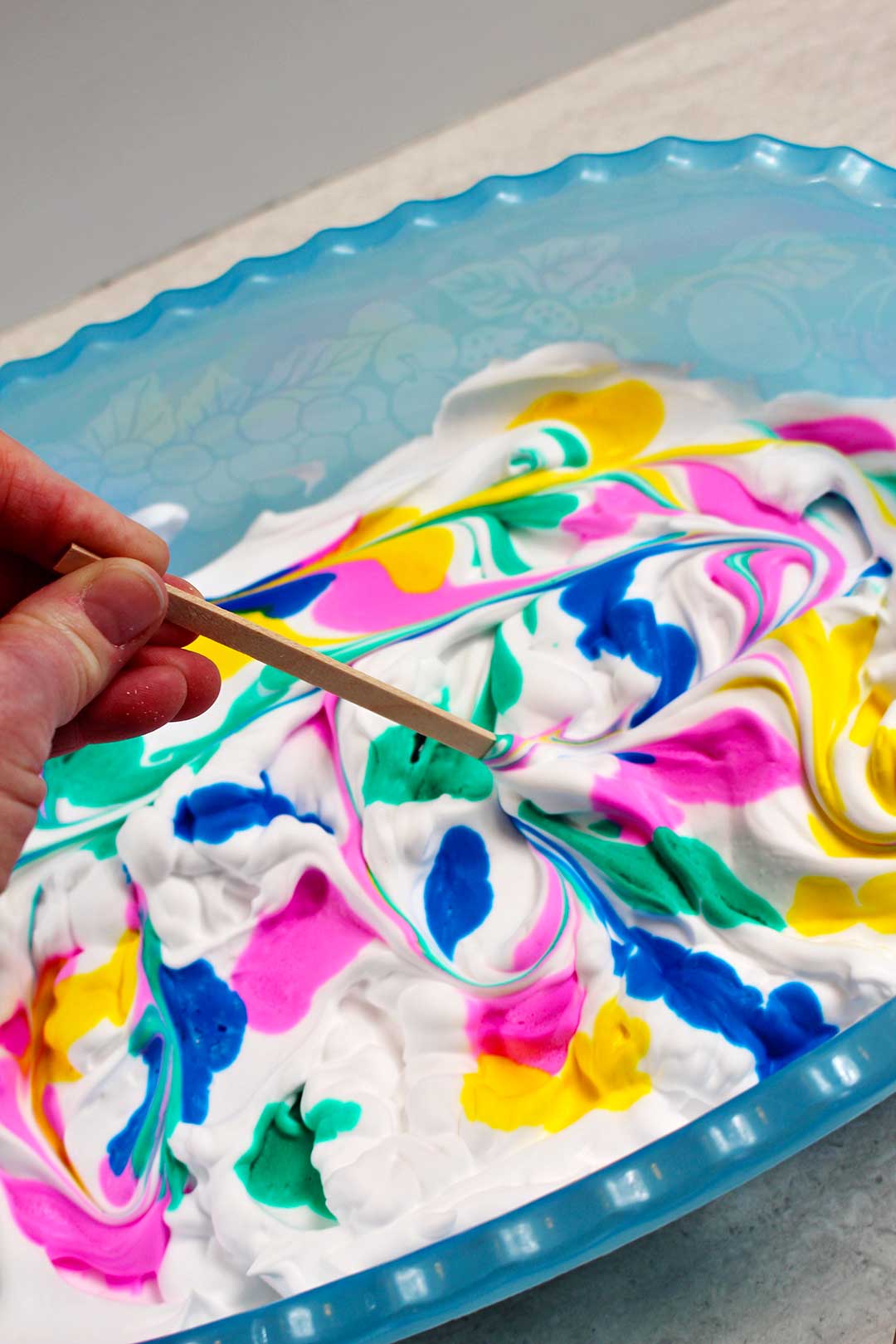 A popsicle stick swirling pink, green, yellow and blue dye in a tub of shaving cream.