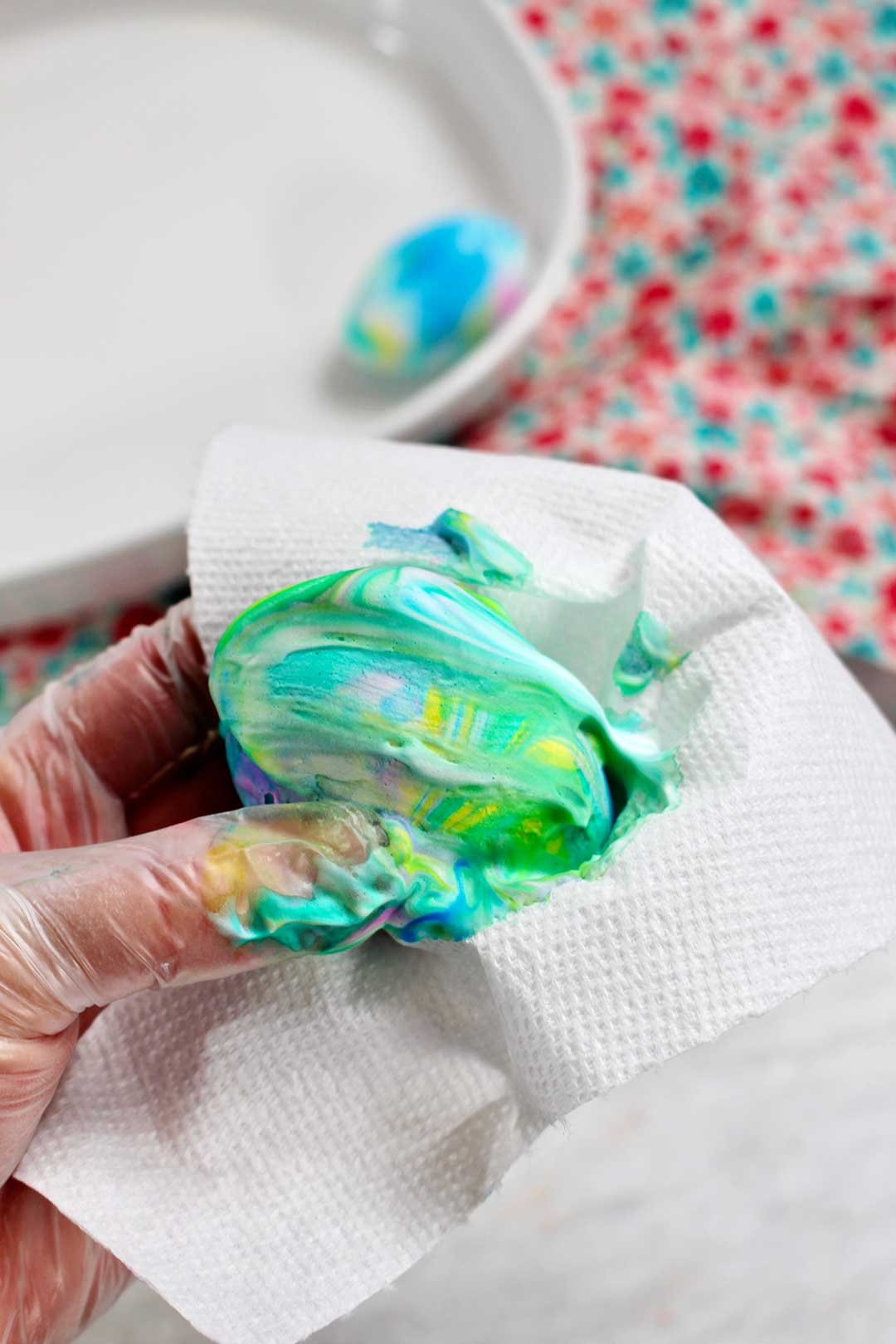 An egg covered in yellow, green, blue, and pink swirled shaving cream, being wiped off by a paper towel.