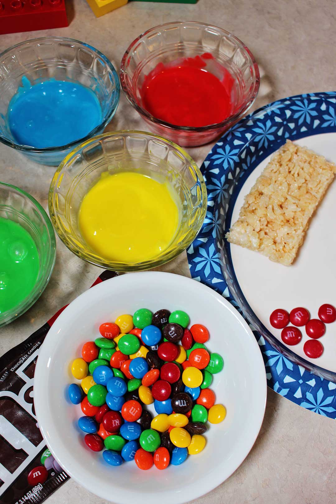 Yellow, blue, red, and green frosting near a bowl of M&Ms and a rice crispy treat on a plate.