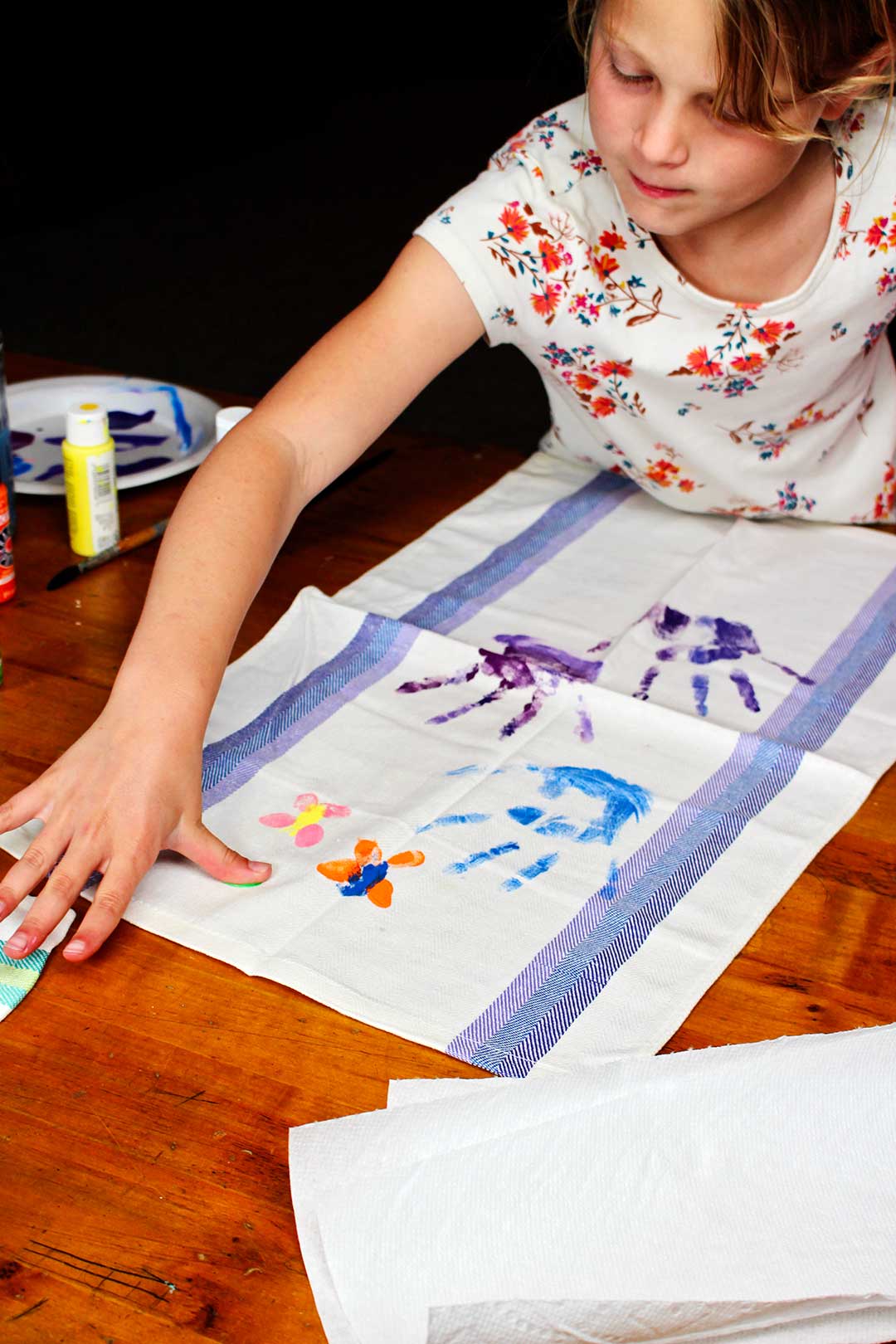A child leaving a green thumbprint on a painted dish towel with handprints.