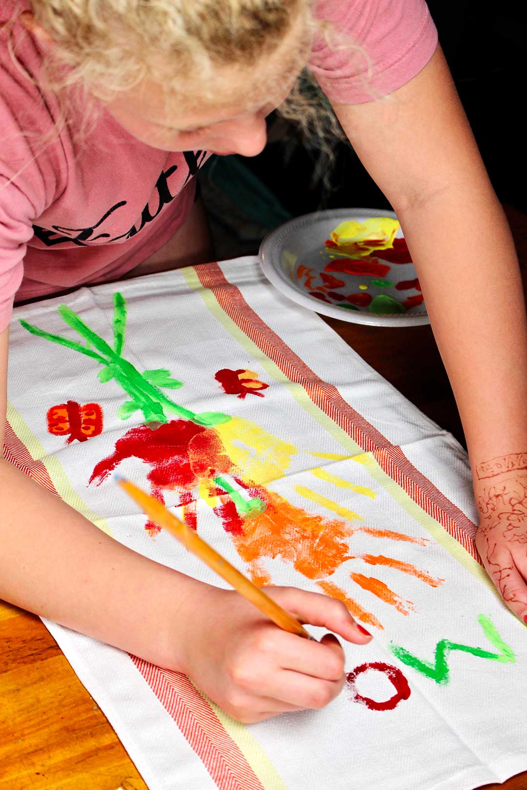 A child painting the word "Mom" on a handprint flower dish towel.