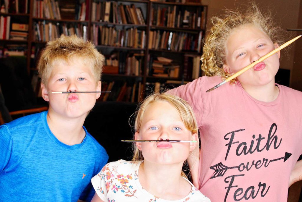 Three kids holding paintbrushes under their noses to create silly mustaches.