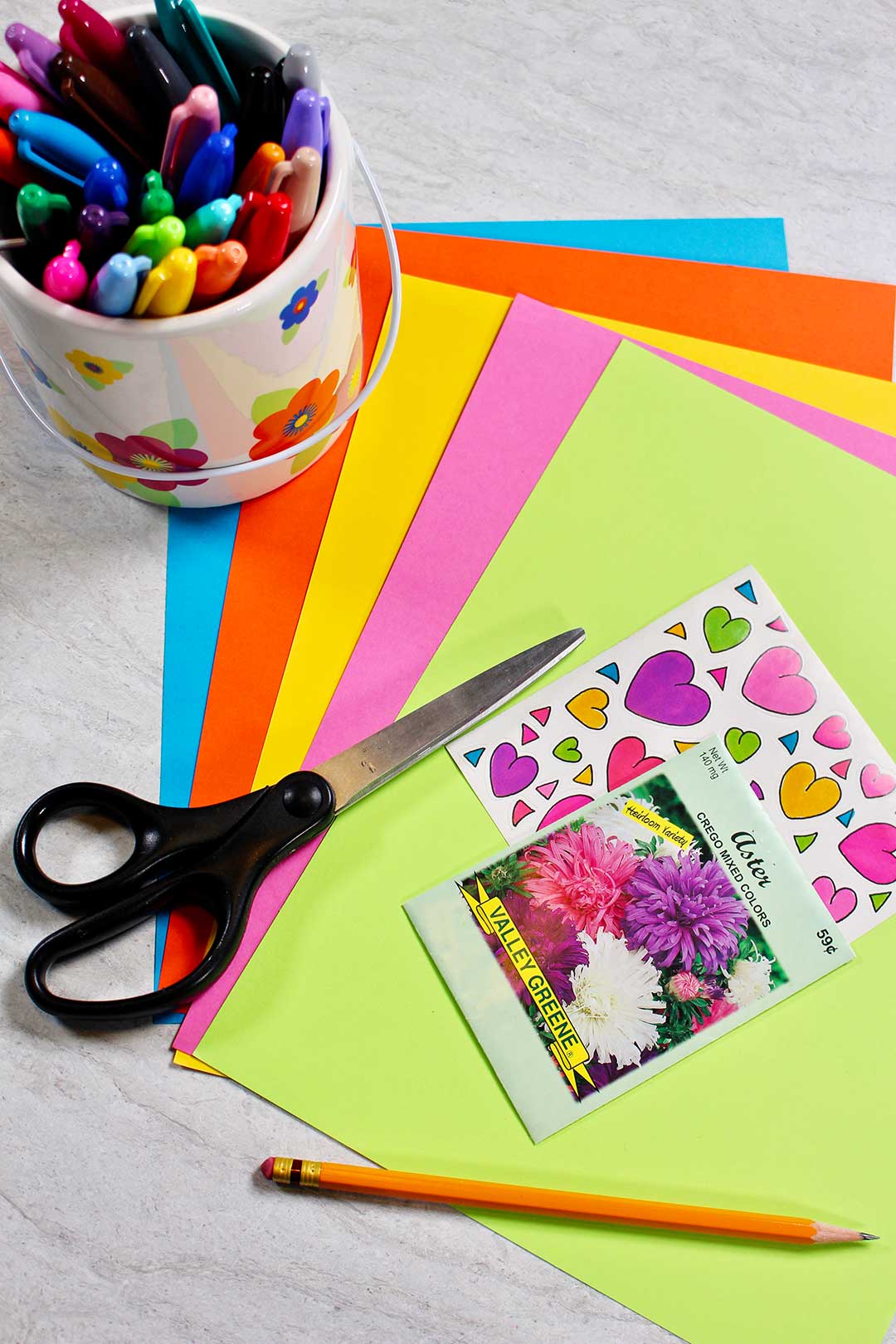 Colorful cardstock paper, flower seeds, heart stickers, a pair of scissors, colorful markers, and a pencil.