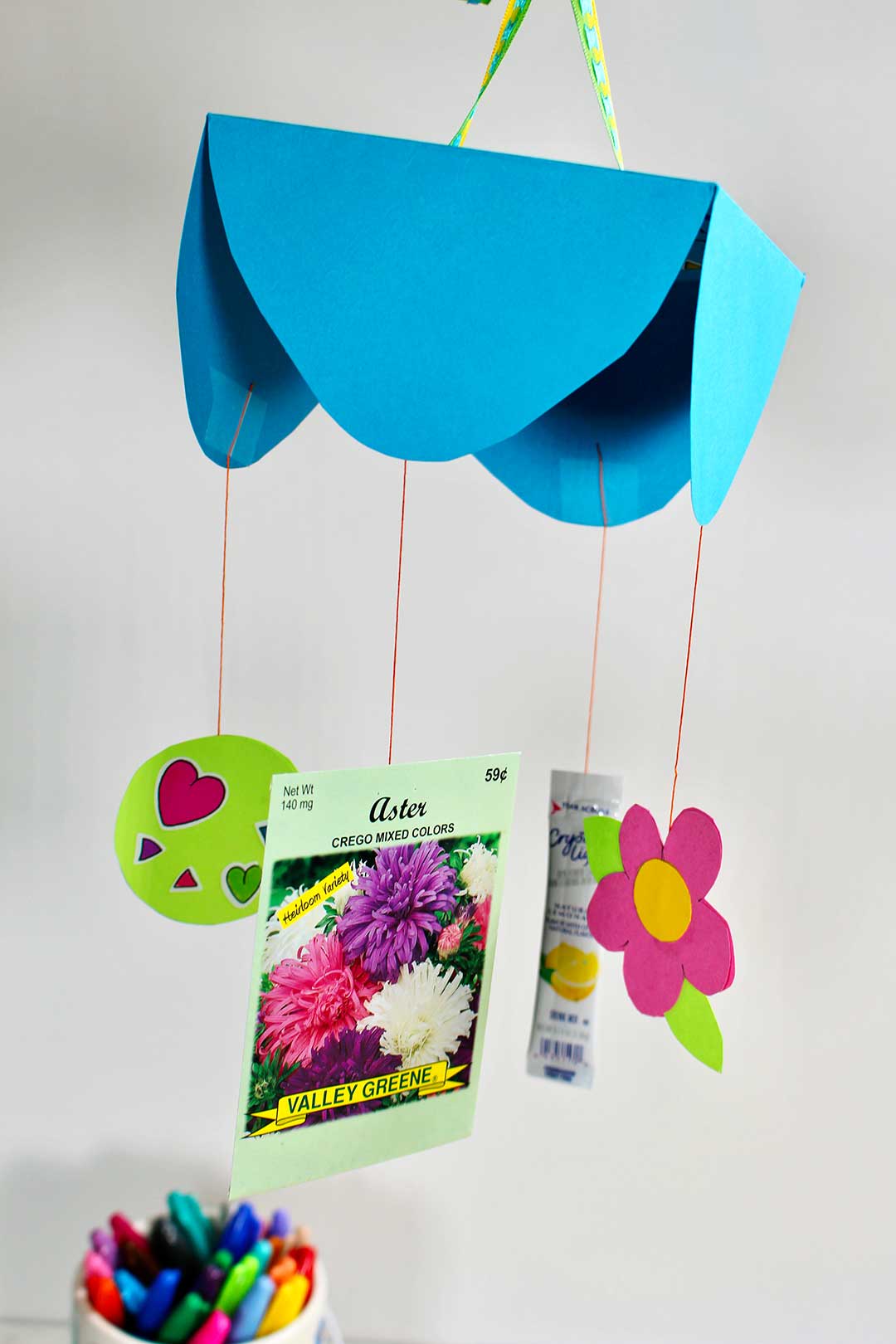 A DIY Mother's Day Card Mobile with stickers, a flower, flower seed packet, and drink packet hanging from strings.