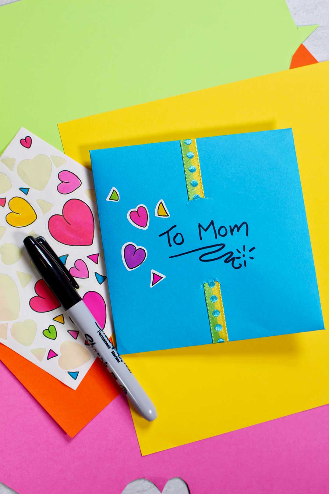 A homemade blue envelope with heart stickers addressed to Mom, ontop a pile of colorful paper.