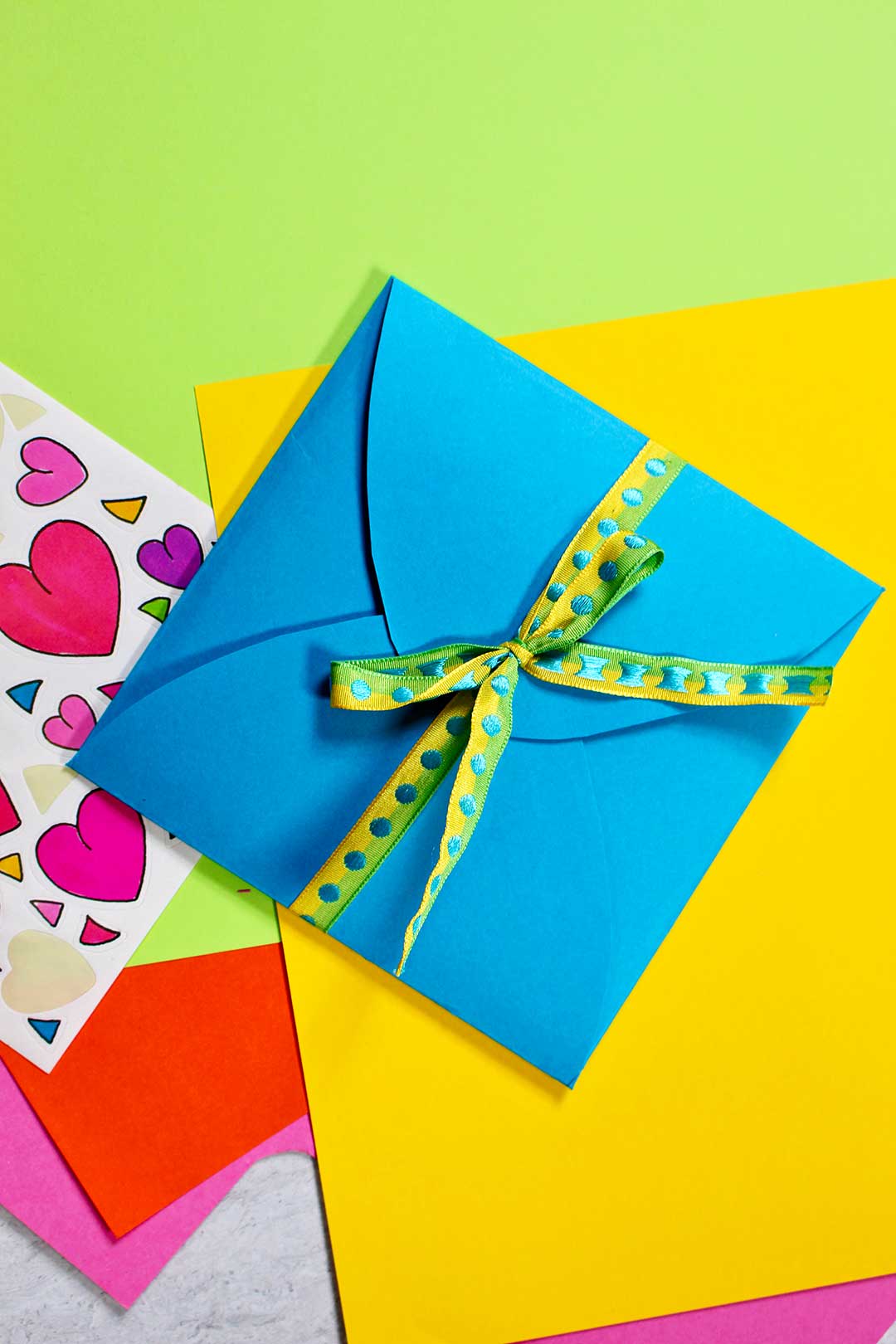 A blue homemade envelope tied with green and yellow ribbon on top a pile of colorful paper and heart stickers.