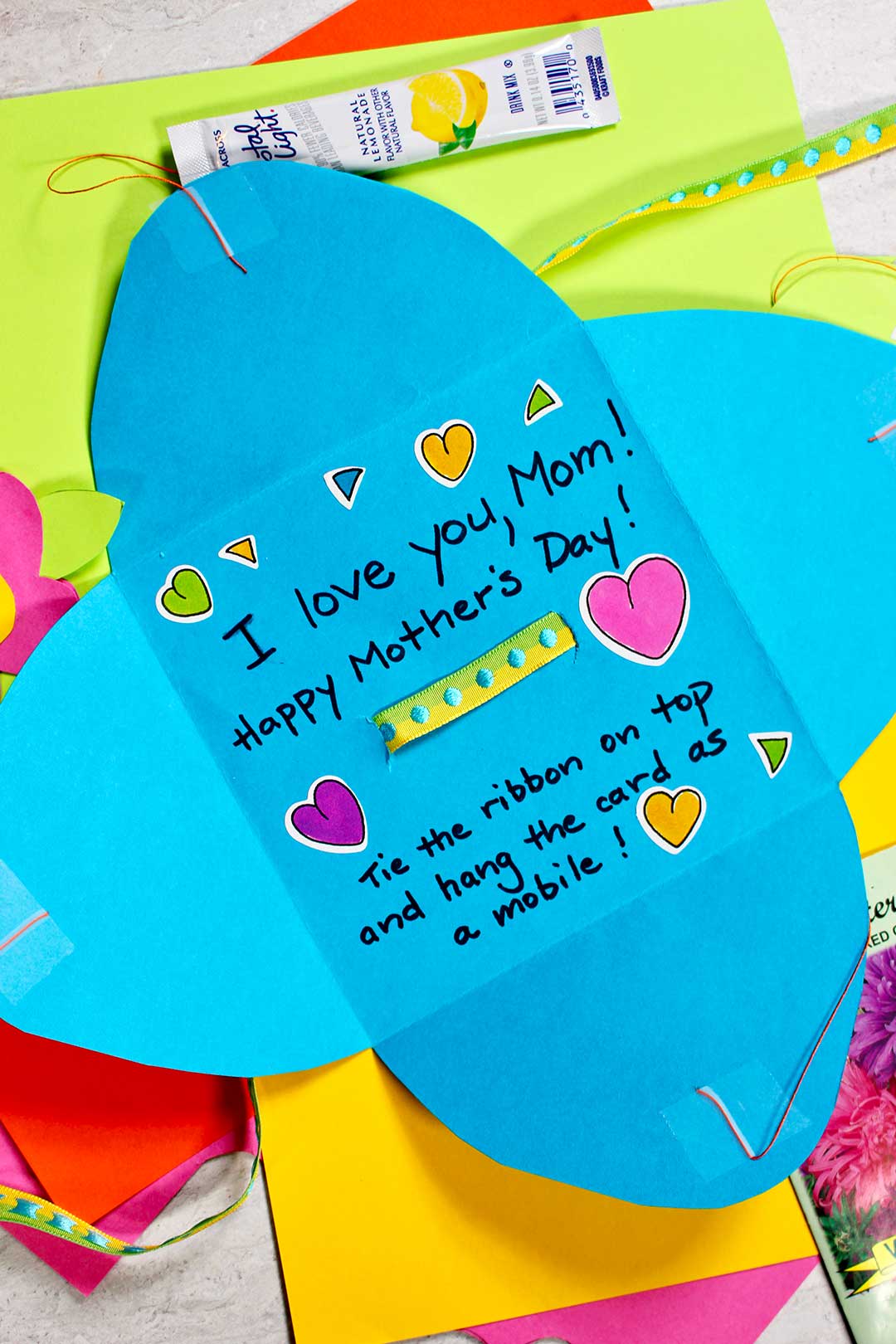 A blue homemade card addressed to Mom for Mother's Day with heart stickers on a pile of colorful paper.
