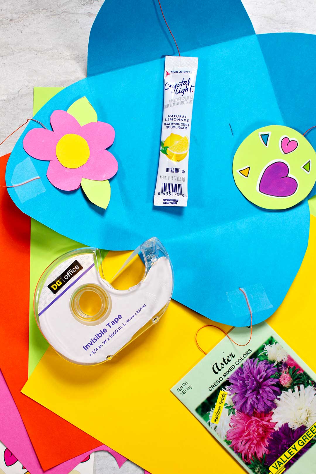 A paper flower, flower seed packet, heart stickers, and a drink packet attached to a blue card with tape.