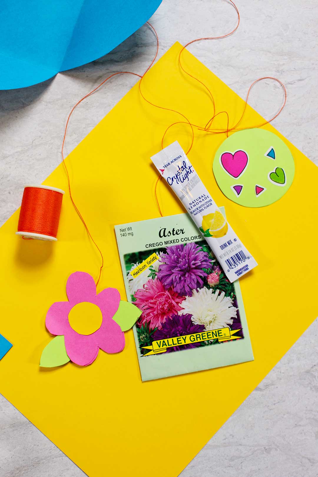 A drink packet, paper flower, flower seed packet, and heart stickers attached to red string.