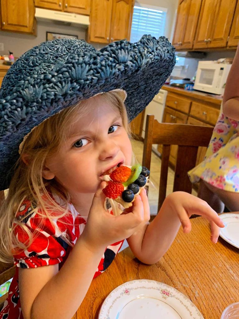 A child in a floral dress and blue hat taking a bite from some fruit pizza.