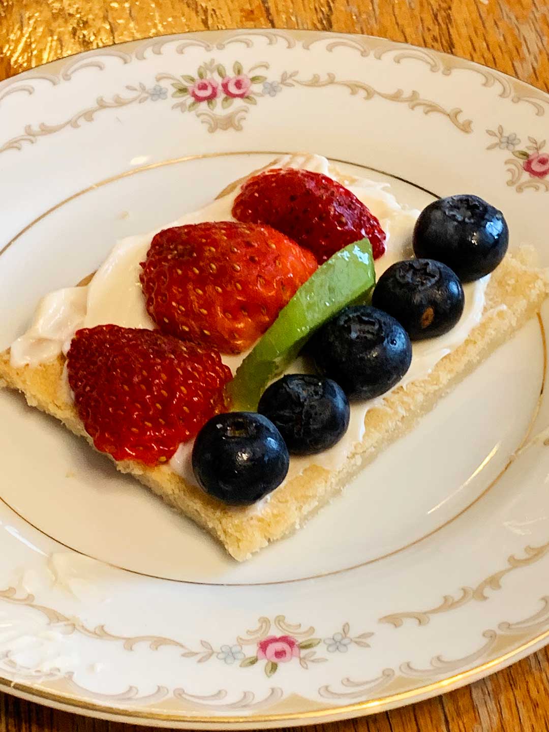 A piece of fruit pizza with frosting, strawberries, blueberries, and kiwi on a decorative plate..