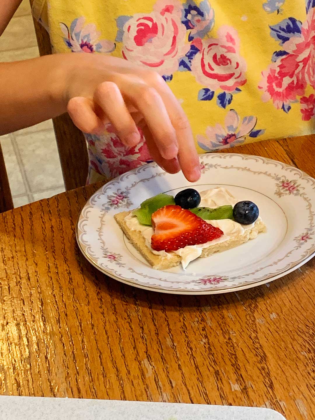 A child decorating a piece of fruit pizza with frosting, strawberries, blueberries, and kiwi on a decorative plate.