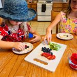 Two children in floral dresses and hats decorating pieces of fruit pizza with frosting, strawberries, blueberries, and kiwi on a decorative plates.