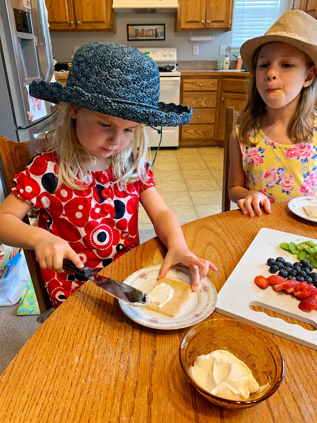 Two children in floral dresses and hats decorating pieces of fruit pizza with frosting, strawberries, blueberries, and kiwi on a decorative plates.