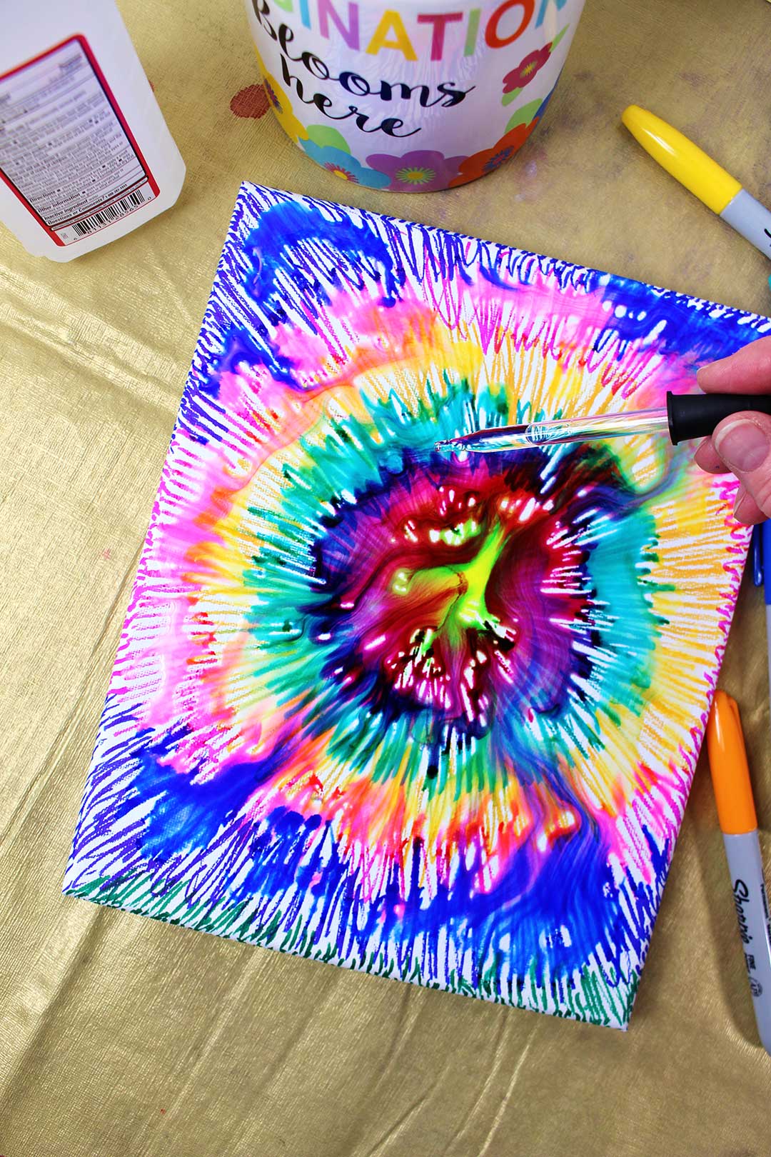 An eye dropper dripping rubbing alcohol over a canvas colored with sharpies in a tie dye pattern.