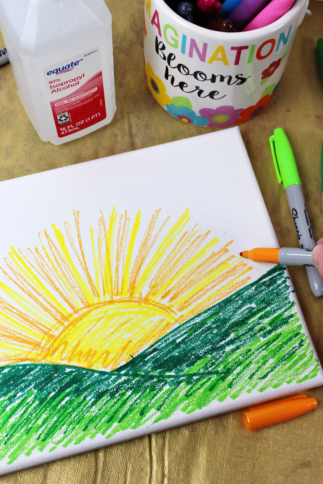 An orange sharpie marker coloring a sunset on canvas, surrounded by a bottle of isopropyl alcohol and sharpie markers.