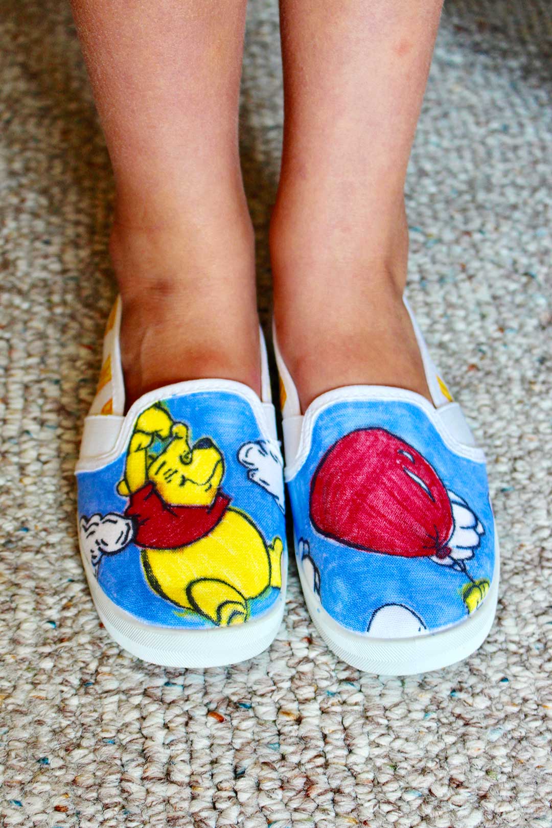 A child wearing a pair of canvas shoes decorated with a marker colored picture of Winnie the Pooh and a balloon.