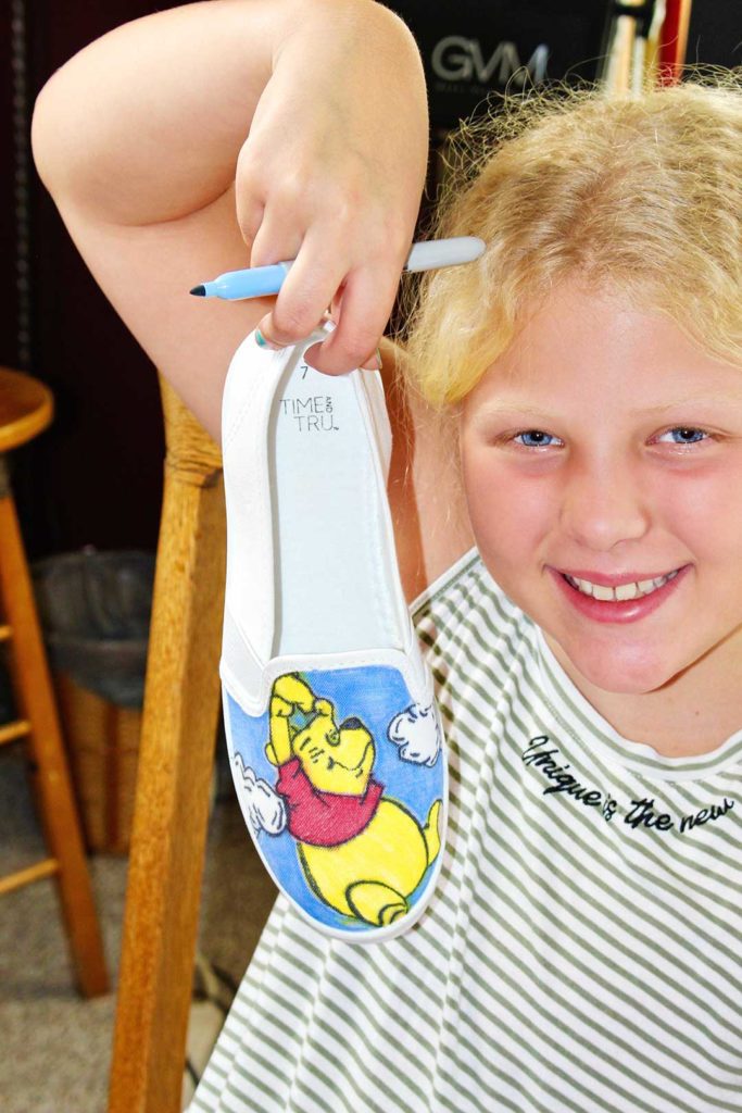A child holding up a white canvas shoe with a colored picture of Winnie the Pooh on the toe.