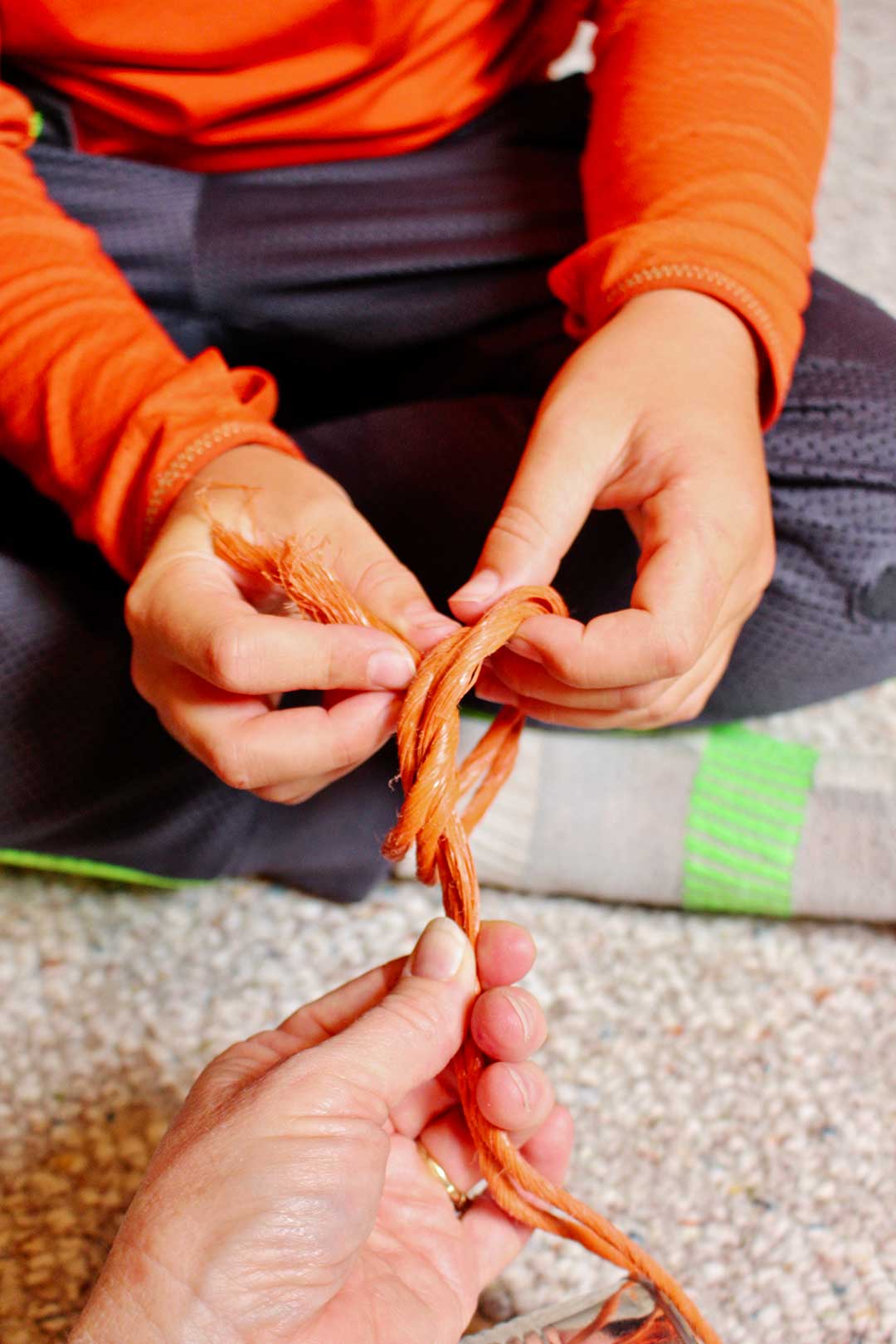 An adult helping a child tie knots in a loop of large orange twine.