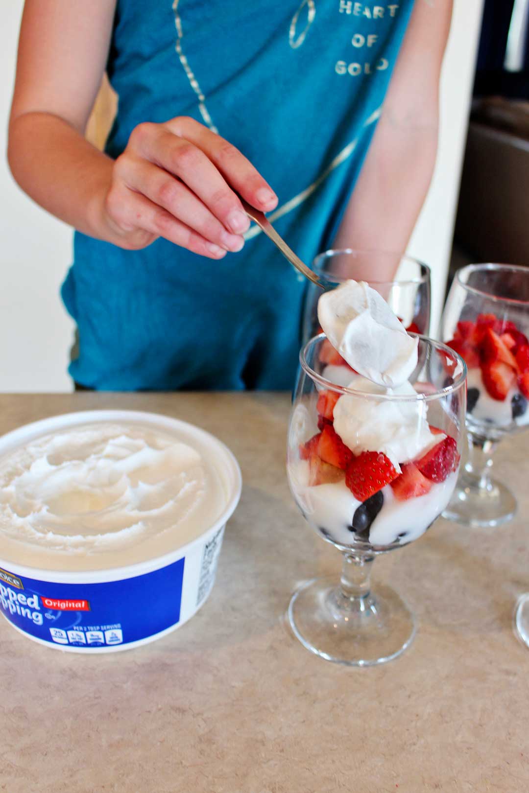 A child adding a scoop of whipped cream to a cup of red and blue fruit.