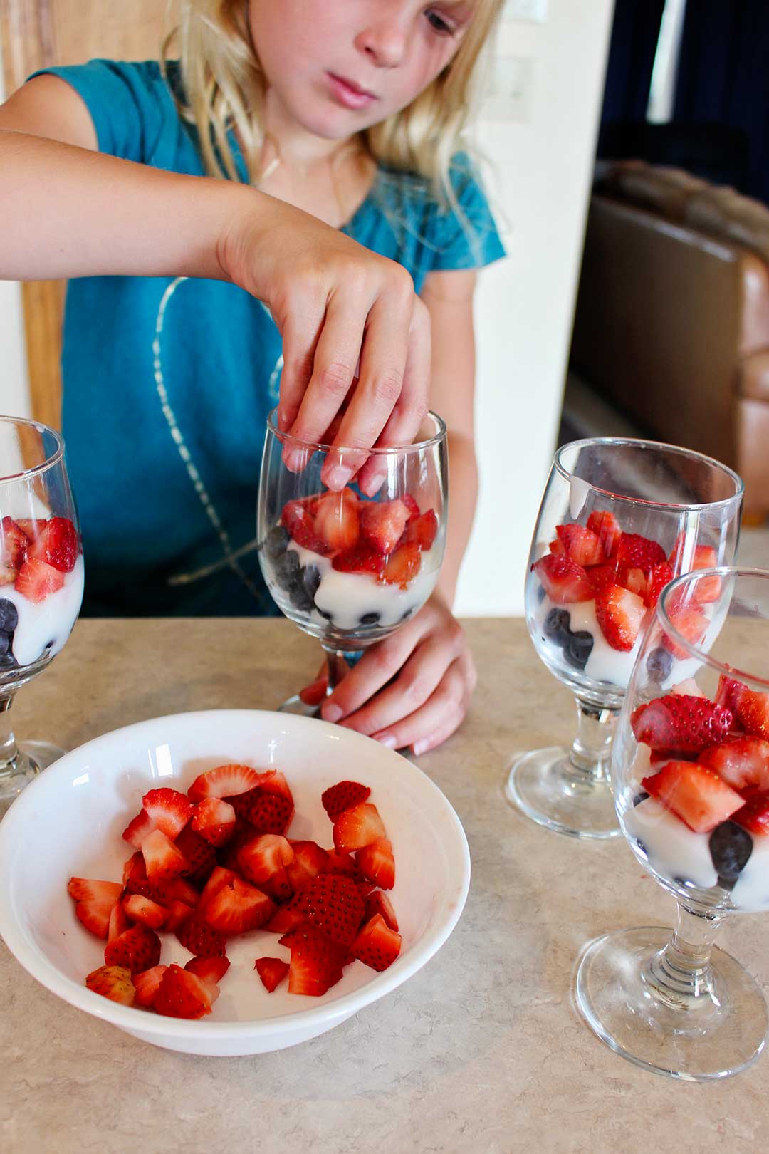 A child adding sliced strawberries to glass cups with blueberries and yogurt.