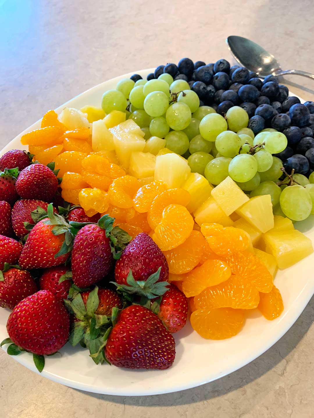 A platter of rainbow fruit with strawberries, oranges, pineapple, green grapes, and blueberries.