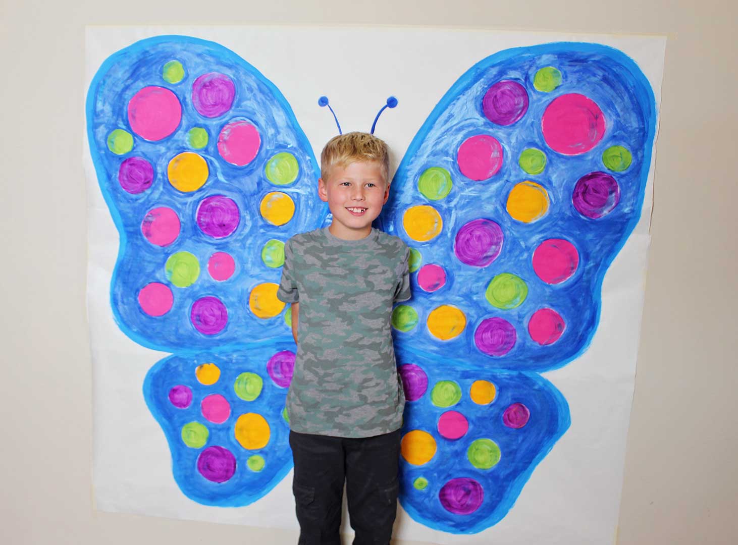 A child standing in front of a painted picture of butterfly wings and antennae.