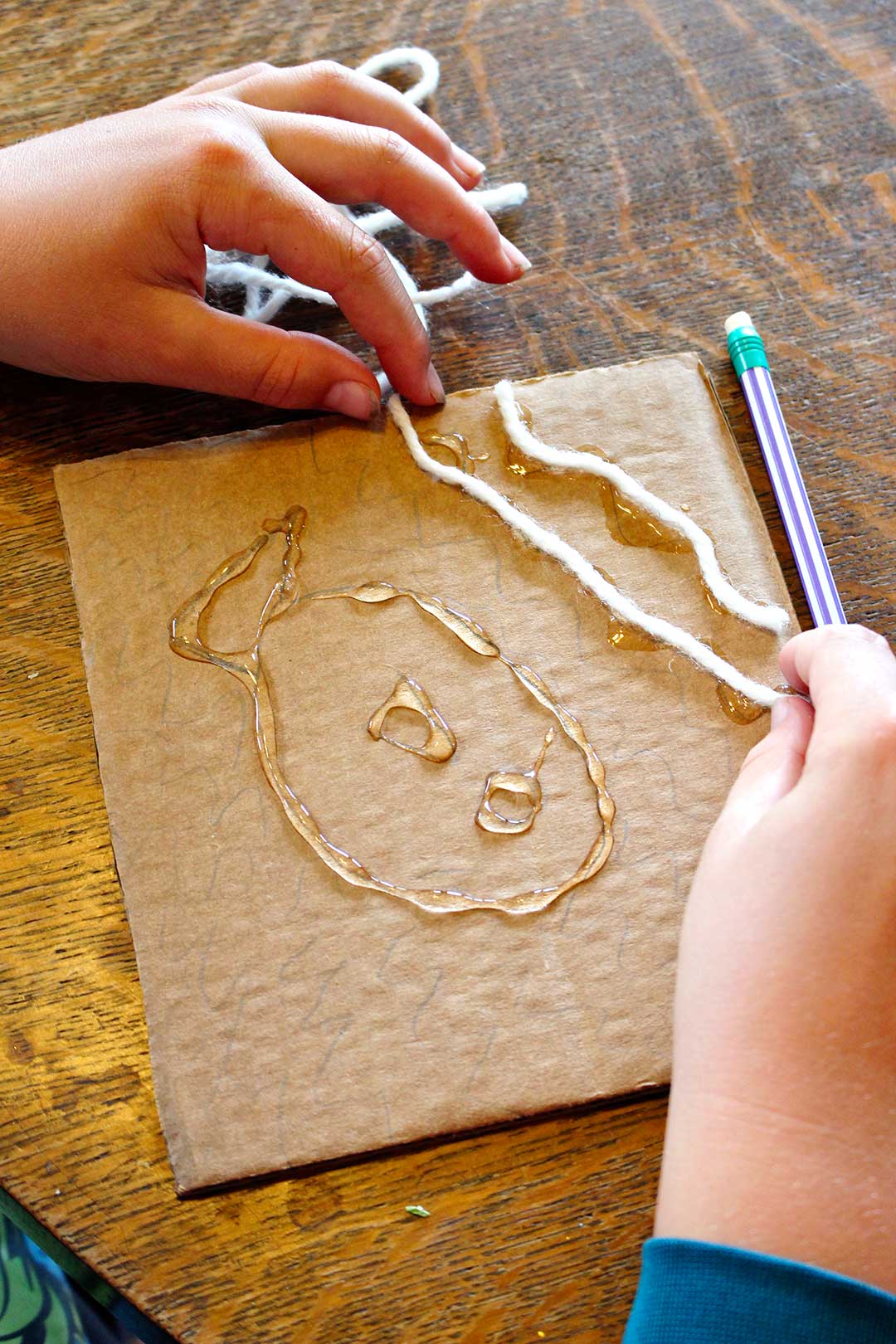 A child gluing a piece of yarn to a square of cardboard in the shape of waves above a fish.