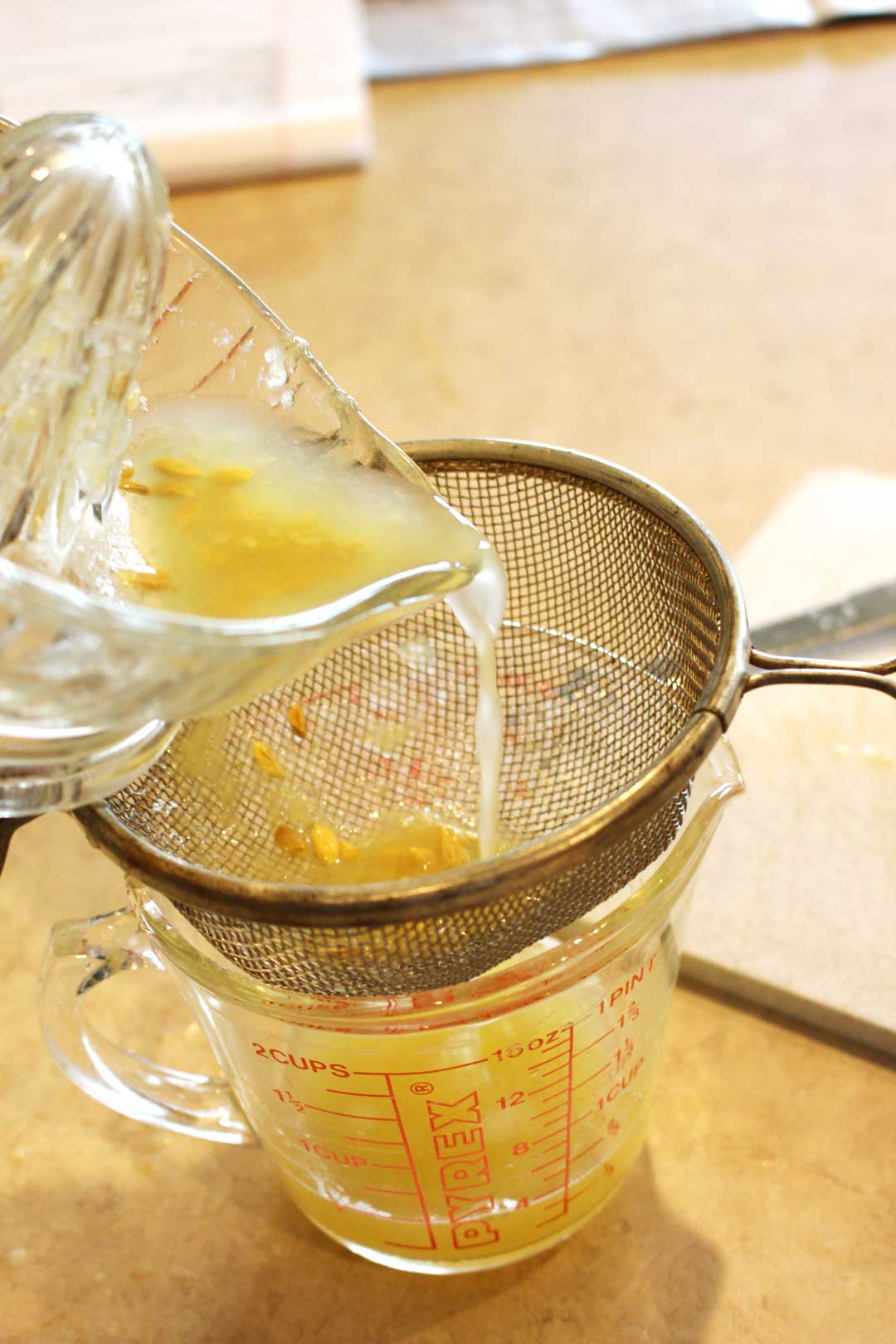 Freshly squeezed lemon juice being poured through a strainer into a glass measuring cup.