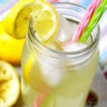 A glass of lemonade in a mason jar with pink and green straws near some lemon slices.
